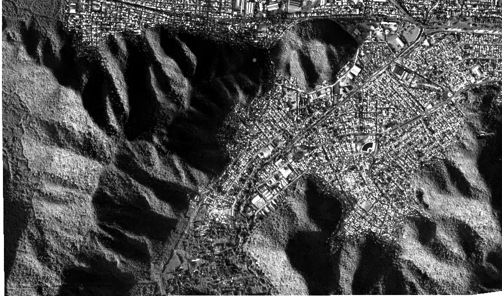 Vantor panchromatic grayscale satellite image of a dense hillside city, with individual buildings, streets, and steep forested ridges visible in fine detail