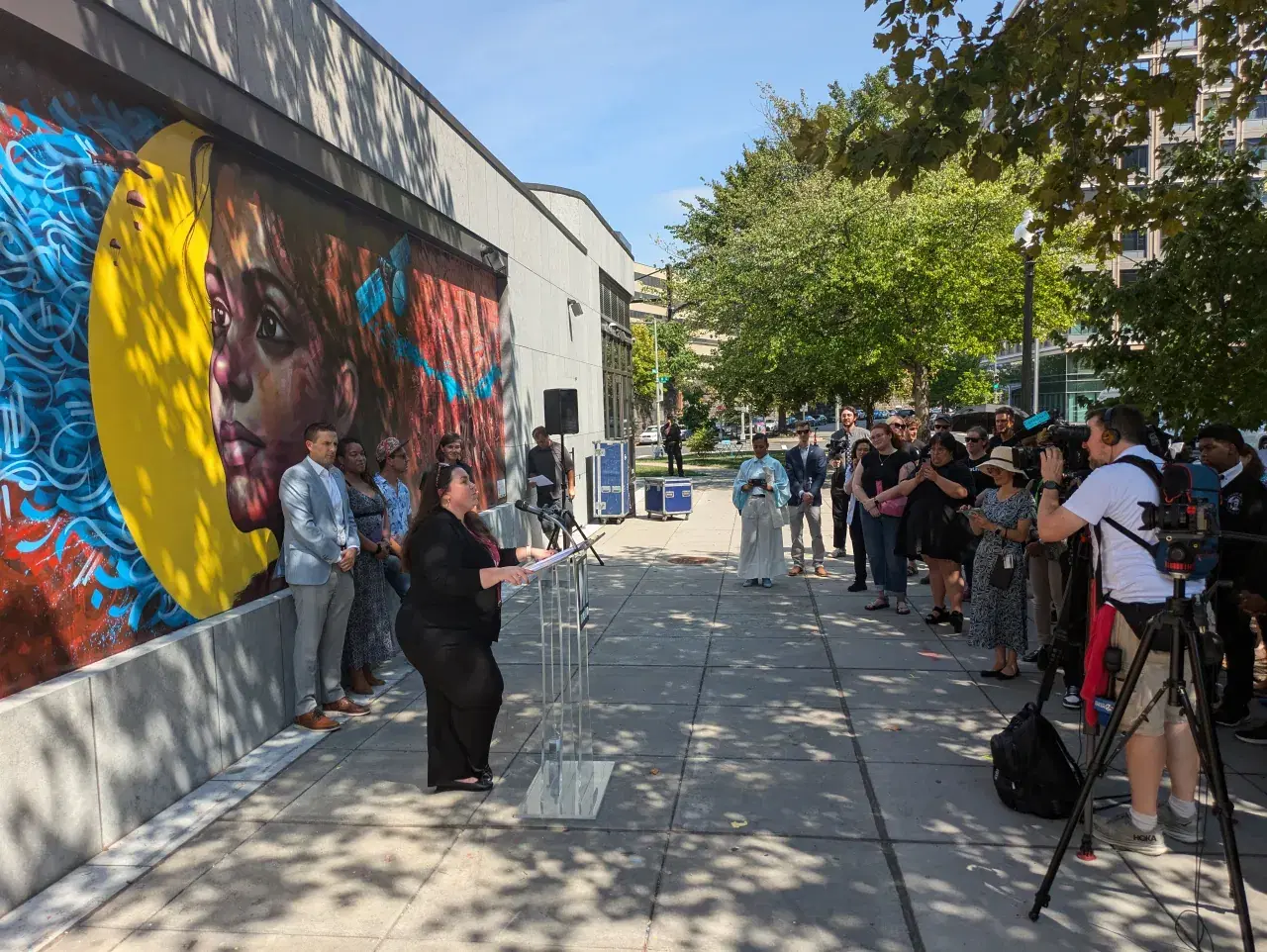 Shanna McClain speaks at a podium in front of the mural on a bright sunny day, with people involved in developing the mural on the left and a large crowd of onlookers and cameramen on the right who are listening to her speech with rapt attention