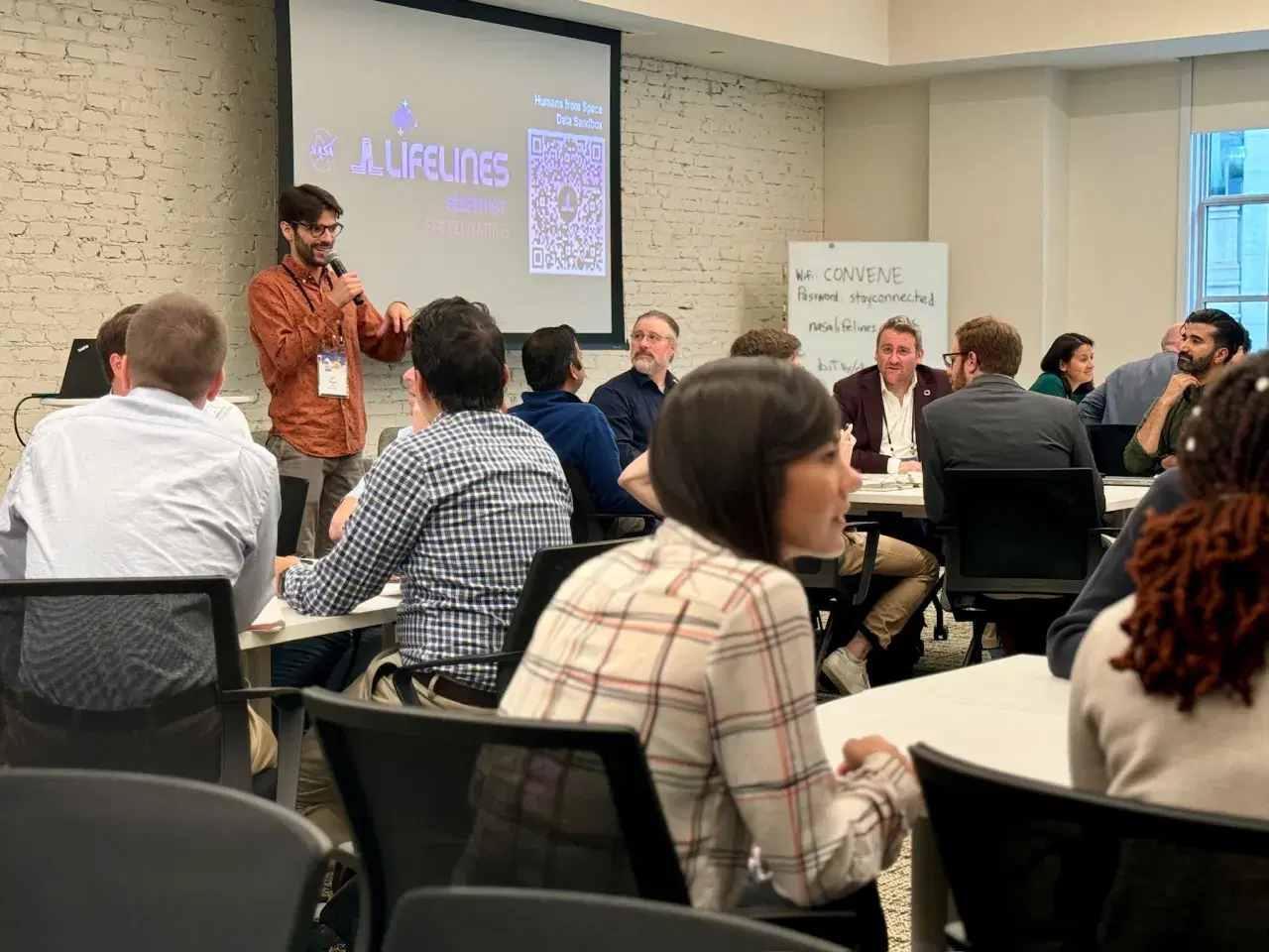 Jeff Pituch stands at the front of a room giving a presentation at the Scientist Speed Dating event. Groups of people sit at tables, eager to participate in this networking event