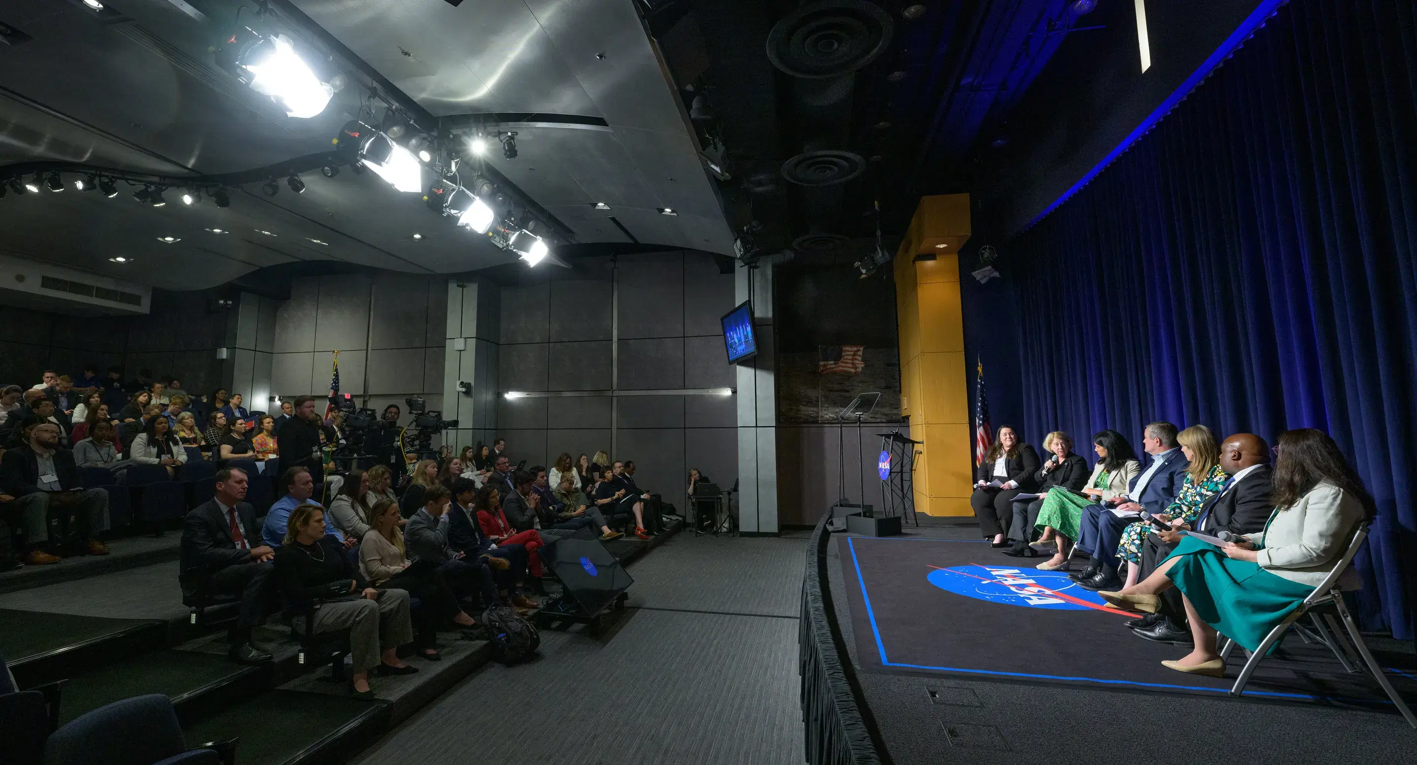 Panelists seated on a stage address a full audience in an auditorium.