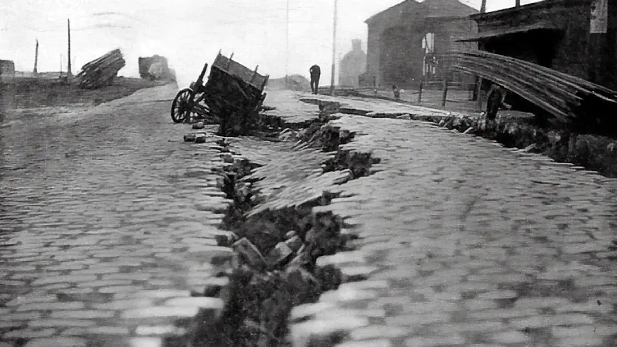 A deep fissure splits a cobblestone street after an earthquake, with an overturned cart tipped into the crack and damaged buildings beyond.