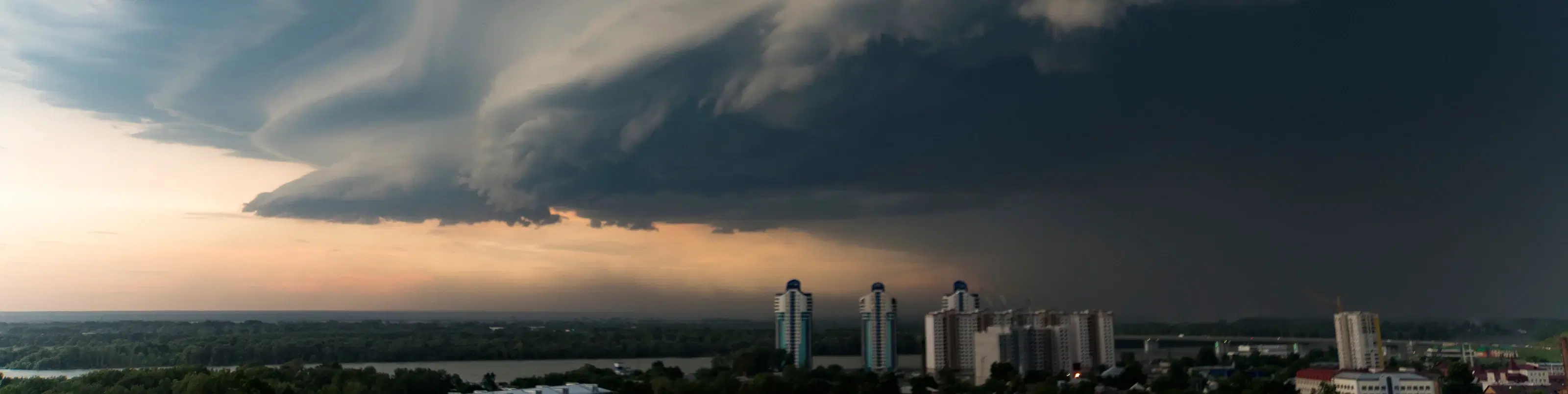 A shelf cloud at the leading edge of an advancing thunderstorm sweeps over a city skyline at dusk.