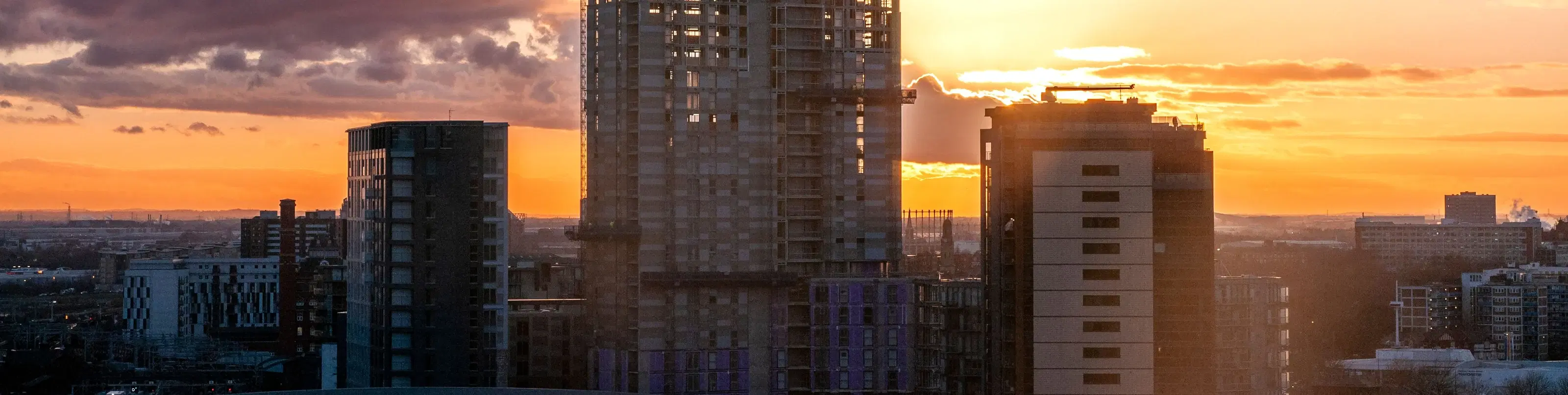 A tower crane rises over high-rise buildings under construction, silhouetted against a city skyline at sunset.