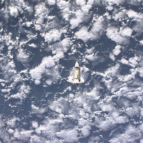 Captured from a distance, the Space Shuttle Endeavour is barely visible; its predominantly white chassis camouflages it against the vast layer of Earth's clouds in the background.