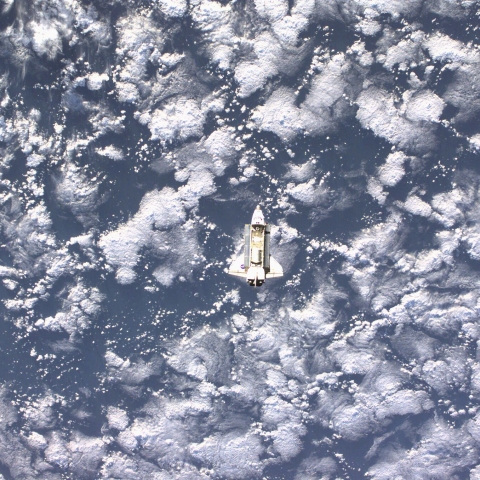 Captured from a distance, the Space Shuttle Endeavour is barely visible; its predominantly white chassis camouflages it against the vast layer of Earth's clouds in the background.